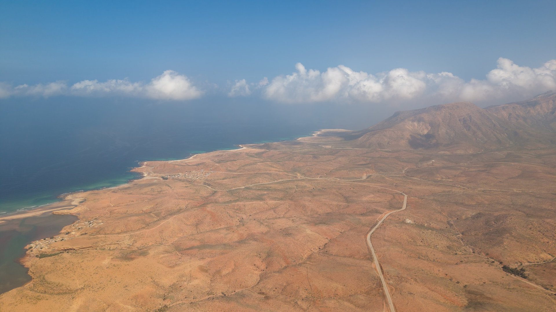 Aerial view of arid coastal landscape with winding road, mountains, and turquoise sea under blue sky