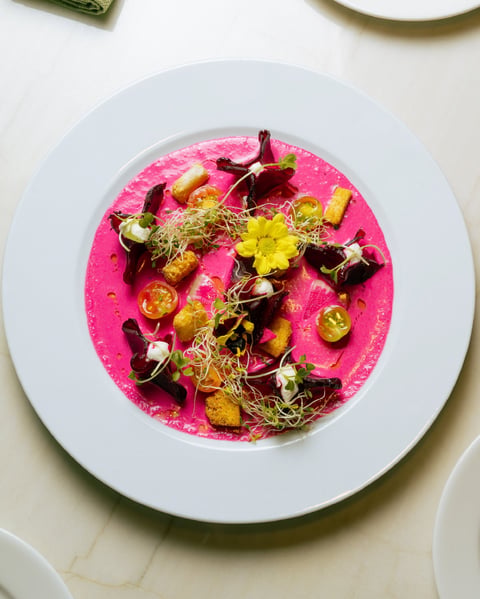 Vibrant pink beet hummus dish topped with roasted vegetables, microgreens, and edible flowers on a white plate