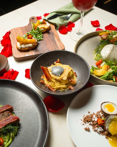 Elegant plated dishes and appetizers arranged on white table with rose petals, including sliced steak, seafood preparations, and a glass of red wine