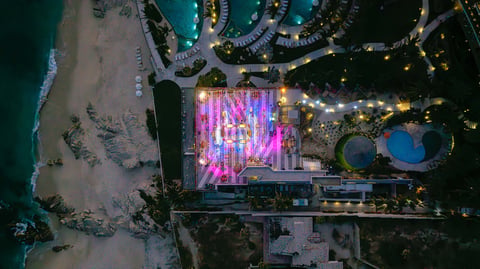 Aerial night view of beachfront venue with pink illuminated stage, surrounded by parking and residential developments along the coastline