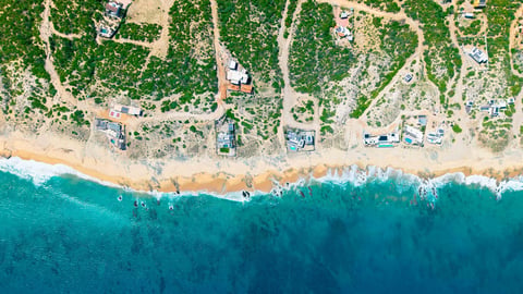 Aerial view of a coastal beach with turquoise water, sandy shoreline, residential properties, and forested areas inland