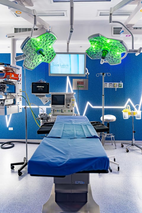 Modern surgical operating room with blue walls, bright green overhead surgical lights, and medical equipment surrounding a patient bed