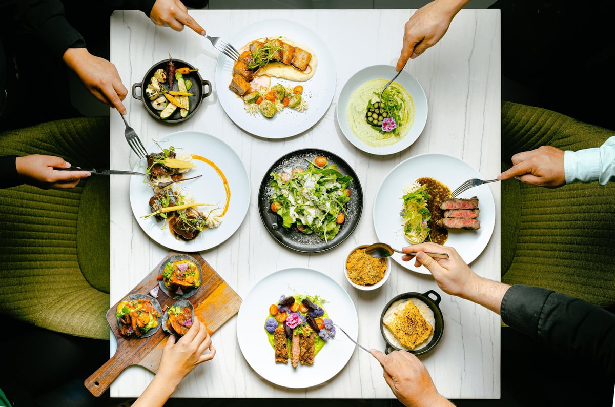 Overhead view of multiple hands sharing various green vegetable dishes and grilled food on white plates and wooden boards around a white table.